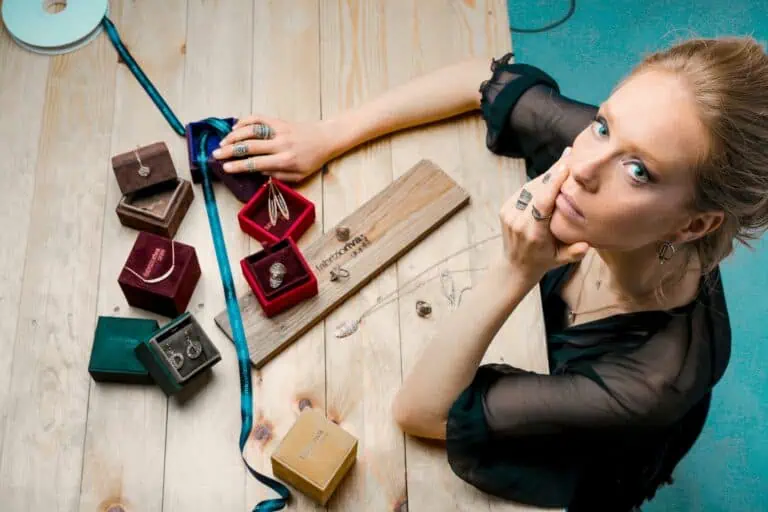 From above serious businesswoman looking up at camera while sitting at wooden table with small jewelry boxes with bijouterie and decorating with ribbon as present