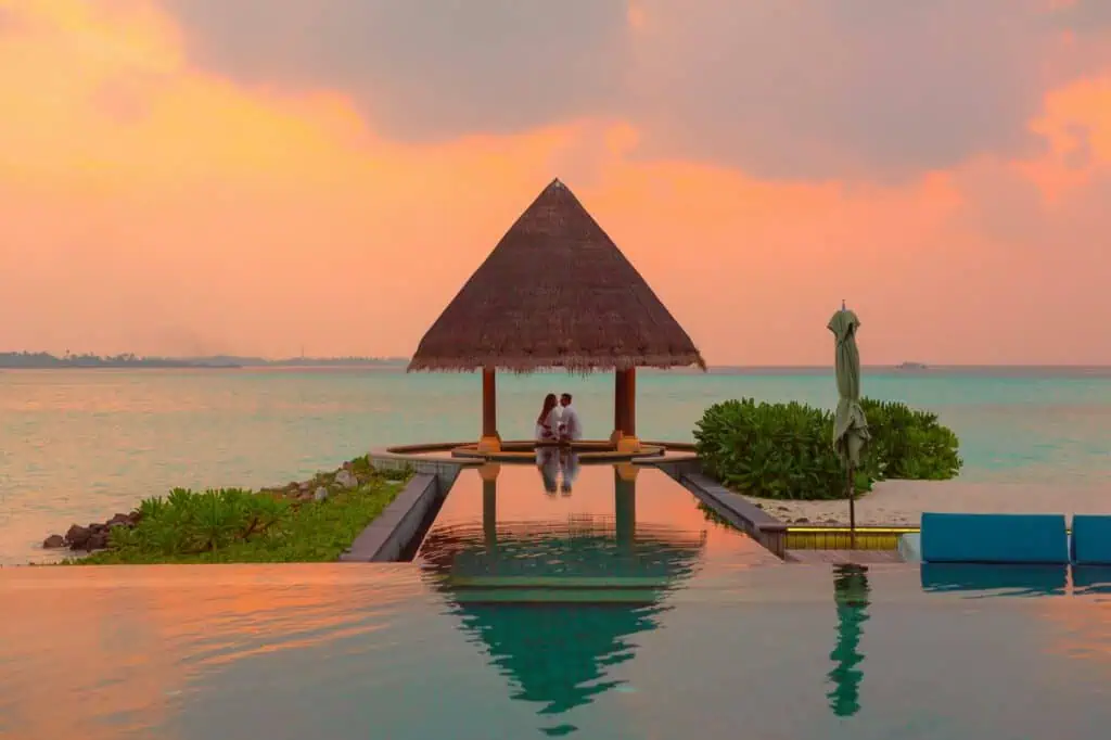 A couple enjoying a romantic moment by the sea and pool under a beach cabana during sunset.