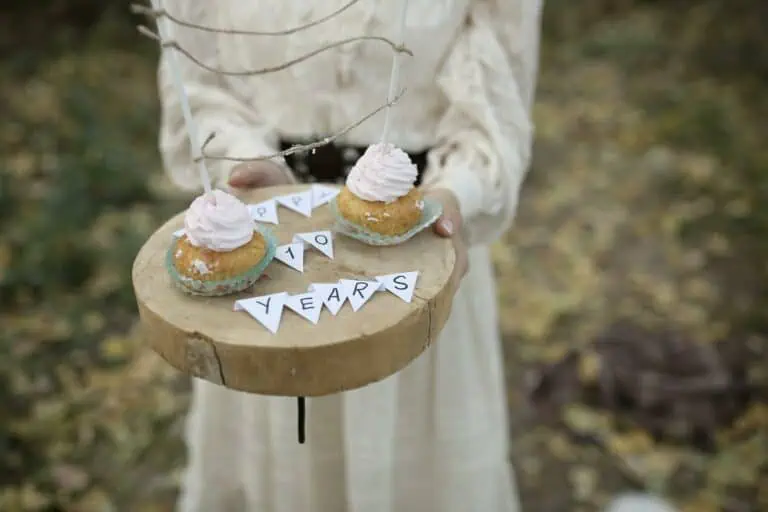 Crop anonymous girl with tasty cupcakes on wooden tray with Happy 10 Years inscription standing on grassy lawn during birthday celebration on blurred background