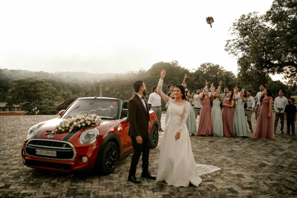 A bride tosses her bouquet at a lively outdoor wedding celebration with guests and a decorated car.